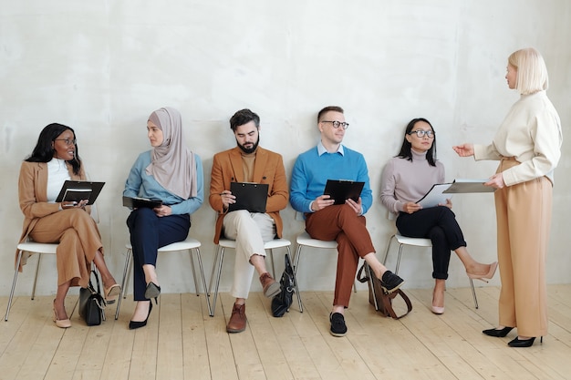 Gestionnaire de recrutement aux cheveux blonds debout devant des candidats à un emploi interracial assis en rang et expliquant le processus d'entretien d'embauche