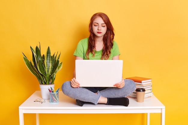 Gingembre jeune étudiante assise sur une table, utilisant un ordinateur portable lors de ses études, regardant l'écran de l'appareil avec une expression faciale concentrée sérieuse, une fille est assise près d'un pot de fleur, pile de livres, stylos, tasse.