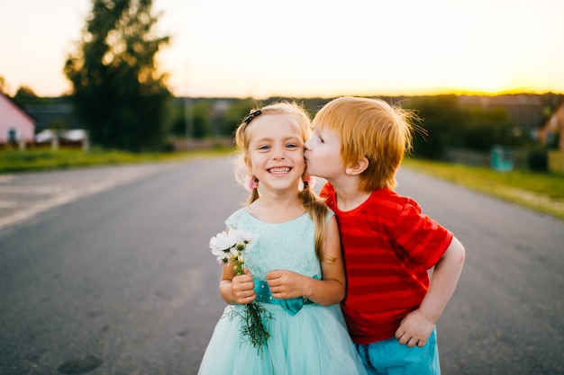 Ginger mec à la peau pâle kising petite fille belle et timide en robe de vacances bleue en plein air sur la route dans la campagne à seunset sur fond abstrait. Automne en terrain rural. Drôles enfants sincères.