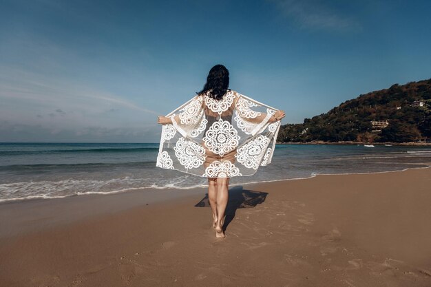 Girlie aux cheveux noirs debout avec son dos à la caméra et a ouvert son paréo comme des ailes de papillon sur une plage contre une mer transparente