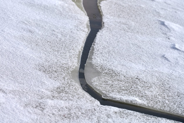 Glace fissurée de rivière gelée avec de la neige blanche sur le dessus et de l'eau bleue en dessous. Fond de texture de glace, gros plan.