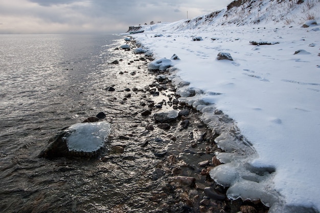 Glace gelée sur des pierres dans l'eau près du rivage en hiver. Temps nuageux. La lumière réfléchie scintille sur l'eau. Neige sur le rivage.