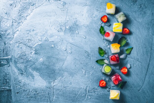 Glaçons avec fruits et glace brisée sur une surface bleu pierre avec des feuilles de menthe et des fruits frais. Menthe, fraise, cerise, citron, orange. Mise à plat, vue de dessus