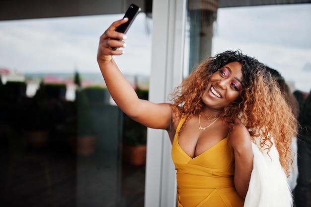 Glamour femme afro-américaine en robe jaune et cape de laine blanche faisant selfie sur téléphone.