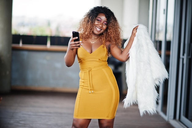 Glamour femme afro-américaine en robe jaune et cape de laine blanche posée au balcon avec téléphone portable à portée de main