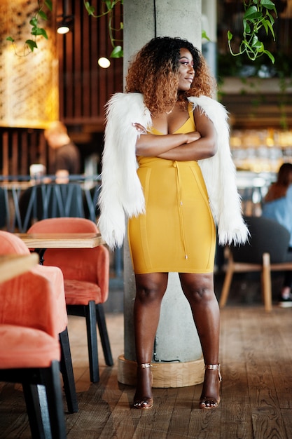 Glamour femme afro-américaine en robe jaune et cape de laine blanche posée au restaurant près de la colonne.