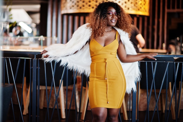 Glamour femme afro-américaine en robe jaune et cape de laine blanche posée au restaurant.