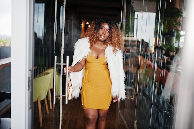 Glamour femme afro-américaine en robe jaune et cape de laine blanche avec sac à main posé à l'entrée de la porte en verre du restaurant.