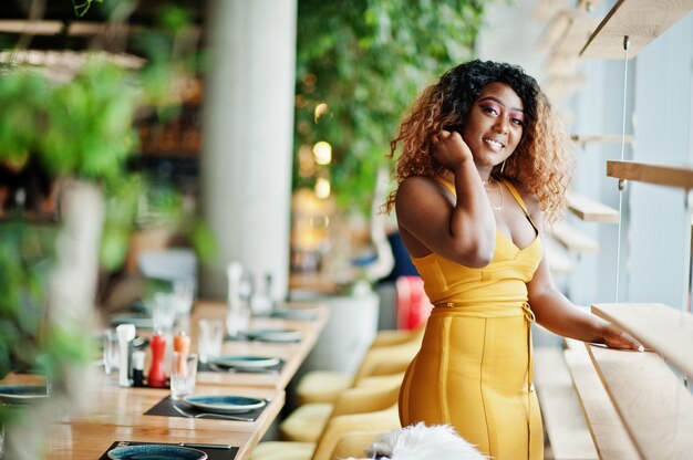 Glamour femme afro-américaine en robe jaune posée au restaurant près de windows.