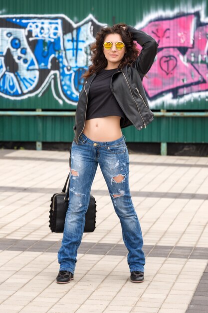 Glamour jeune femme brune en veste de cuir noir et lunettes de soleil s'amuser dans la ville