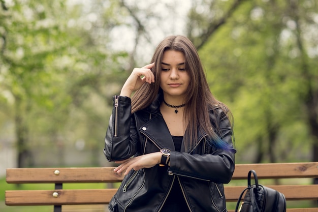 Glamour jeune femme en veste de cuir noir assis dans le parc sur le banc