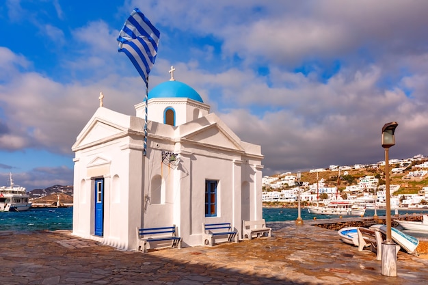 Église chrétienne d'Agios Nikolaos, bâtiment d'église grec typique et grand drapeau grec ondulant dans le vieux port de la ville de Mykonos, Chora, sur l'île de Mykonos, l'île des vents, Grèce