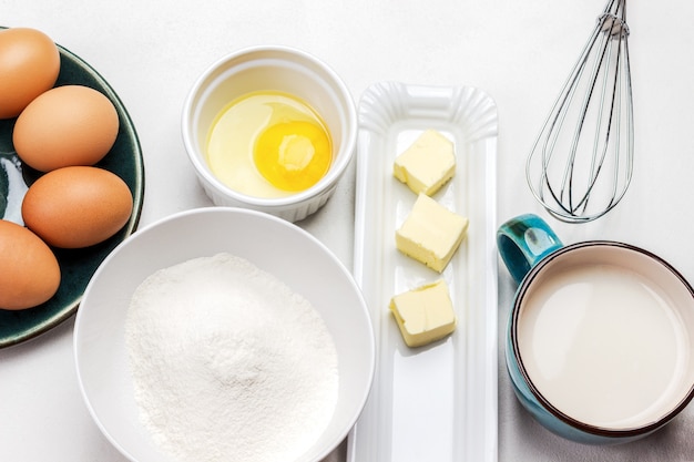 Glor dans un bol, du lait dans une tasse, du beurre sur une assiette et des œufs bruns dans un bol gris sur fond blanc