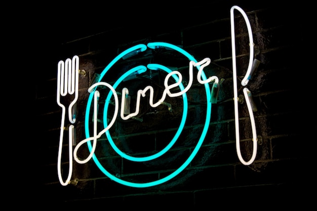 Photo glowing neon inscription dîner et fourchette stylisée, couteau et assiette sur fond de mur de briques sombres. image vintage des tons sombres.