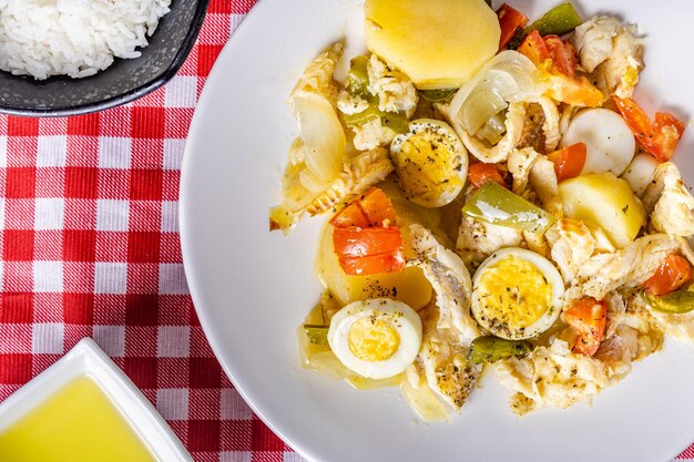 Gomes de sa morue rôtie à l'huile d'olive avec tomates poivrons oignons œufs durs et origan Dans un plat en porcelaine blanche sur une nappe à carreaux avec des couverts riz et huile d'olive