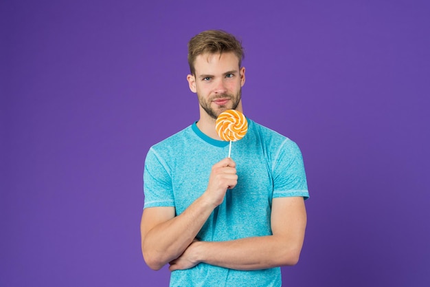 Goût de l'enfance Homme beau macho mange grosse sucette sucrée colorée Homme aux poils aime la sucette Concept de repas de triche Sucre nocif pour la santé Guy tient sucette bonbon fond violet