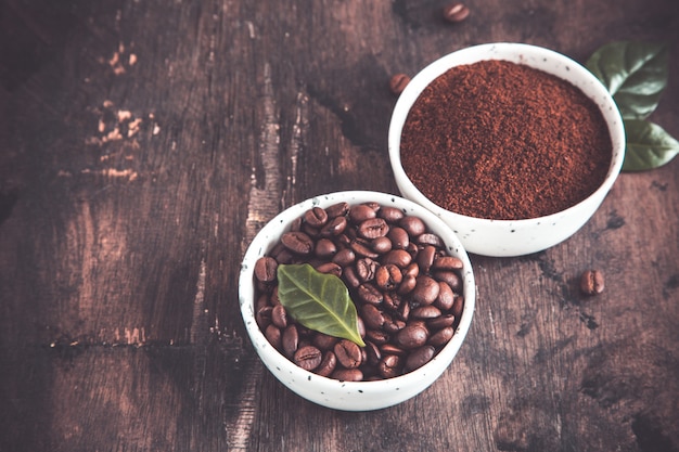 Grains de café et café moulu dans des bols avec des feuilles de caféier sur un fond sombre.