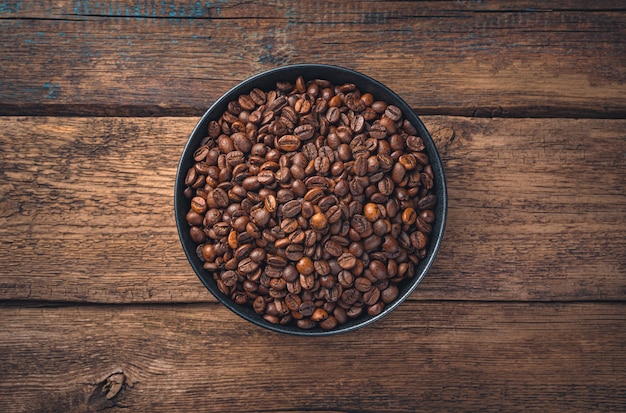 Grains de café dans une assiette en céramique ronde noire sur un mur en bois marron. Vue de dessus.