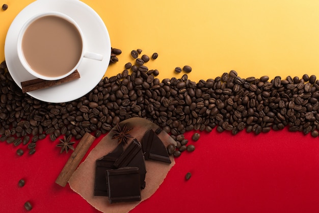 Les grains de café sont dispersés sur un papier rouge et jaune et une tasse blanche, cannelle, anis étoilé, chocolat, fond blanc.