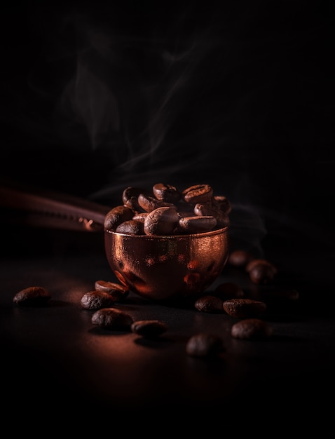 Grains de café torréfiés dans la cuillère en cuivre avec de la fumée sur fond sombre. Concept pour reconnaître l'arôme du café.
