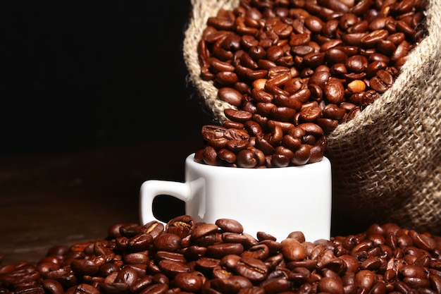 Grains de café torréfiés frais dans un sac en jute, une tasse à café et un moulin sur fond sombre.