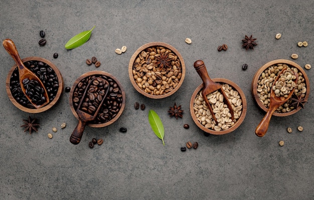 Grains de café torréfiés verts et bruns non torréfiés et foncés dans un bol en bois avec des cuillères installées sur fond de béton foncé