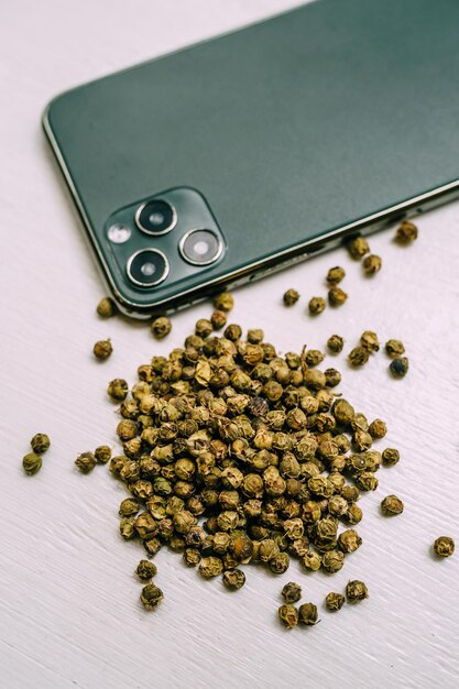 Des grains de poivre vert séchés sont éparpillés sur une table blanche près d'un téléphone portable. Épices et condiments pour la cuisine