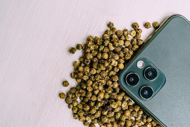 Des grains de poivre vert séchés sont éparpillés sur une table blanche près d'un téléphone portable. Épices et condiments pour la cuisine