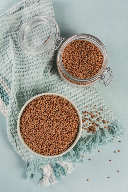 Grains de sarrasin dispersés non cuits biologiques dans un bol et bocal en verre sur fond bleu clair