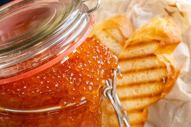 Photo grand caviar rouge dans un bocal, ramasser avec une cuillère, se trouve sur du parchemin, à côté d'une miche de pain baguette