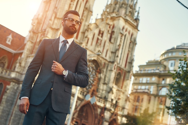 Grand homme d'affaires confiant et élégant en costume complet et lunettes regardant loin en se tenant debout