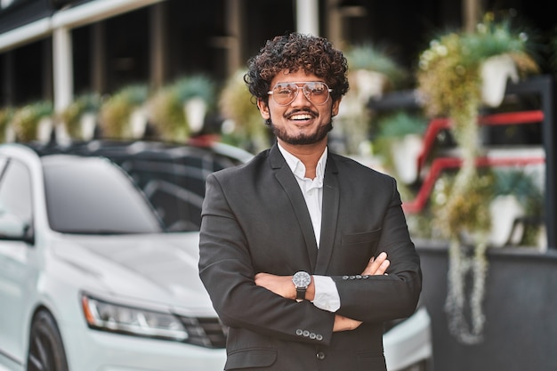 Grand homme d'affaires indien avec des lunettes près de la voiture en costume.