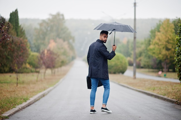 Grand homme à la mode barbe arabe portant un manteau noir avec un parapluie et un sac posé le jour de la pluie