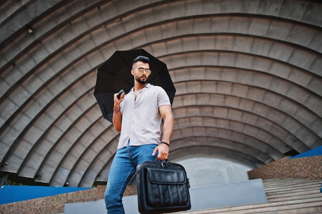 Grand homme à la mode barbe arabe porter sur chemise jeans et lunettes de soleil avec parapluie et sac de cas posés au hall de l'arène