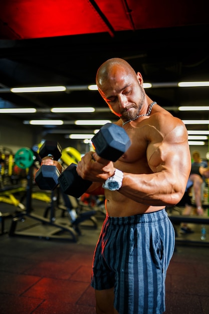 Grand homme musclé avec un torse nu avec des haltères dans les mains. L'homme dans le gymnase secoue les biceps