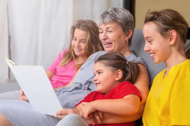 Grand-mère lit un livre à ses petits-enfants Senior et filles sur le canapé en écoutant des contes de fées