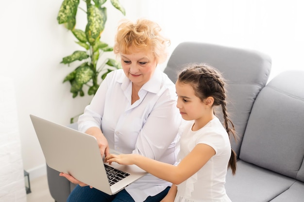 Grand-mère mature aidant l'enfant à faire ses devoirs à la maison. Vieille grand-mère satisfaite aidant sa petite-fille à étudier dans le salon. Petite fille écrivant sur un ordinateur portable avec un professeur principal assis à côté d'elle