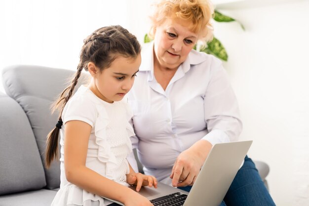 Grand-mère mature aidant l'enfant à faire ses devoirs à la maison. Vieille grand-mère satisfaite aidant sa petite-fille à étudier dans le salon. Petite fille écrivant sur un ordinateur portable avec un professeur principal assis à côté d'elle