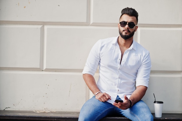 Grand modèle d'homme arabe élégant en chemise blanche, jeans et lunettes de soleil posés dans la rue de la ville et tenir le téléphone portable à portée de main Barbe arabe avec une tasse de café assis et se reposer