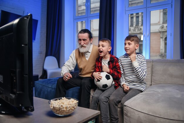 Grand-père barbu senior concentré et sympathique avec ses petits-fils joyeux passant leur temps libre à réviser le match de basket à la télévision