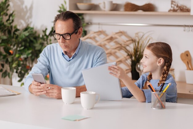 Grand-père concentré occupé et âgé utilisant un smartphone à la maison et ignorant sa petite-fille tout en travaillant et en envoyant des SMS