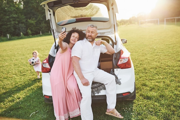 Grand-père Et Grand-mère Sont Assis Ensemble Sur La Nature Près De La Voiture. Ils Font Une Photo De Selfie Et Leur Petite-fille Joue Avec Eux