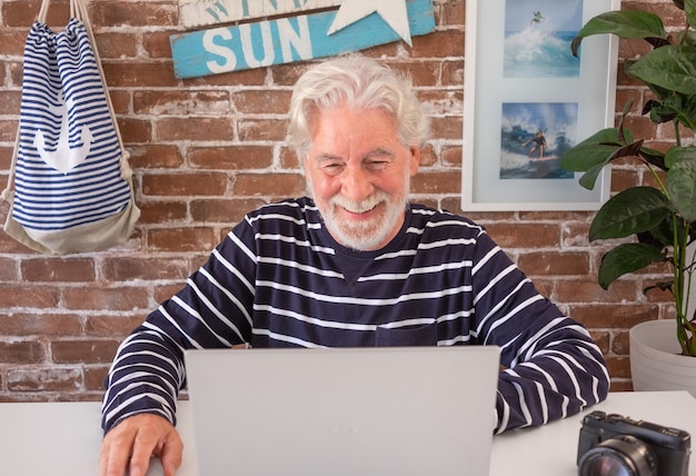 Grand-père marin à la retraite utilisant un ordinateur portable pour passer un appel vidéo à son petit-fils. Heureux homme âgé aux cheveux blancs, profitant de la technologie et du social. Mur de briques en arrière-plan et décorations