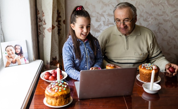 grand-père et petite-fille parlent par liaison vidéo à leurs amis. Table décorée avec des œufs colorés et des gâteaux. Discuter pendant la pandémie COVID et les vacances de Pâques.