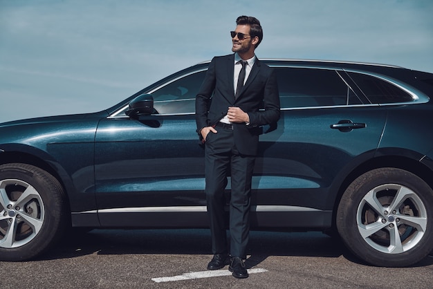 Grand sens du style. Toute la longueur du beau jeune homme d'affaires souriant tout en se tenant près de sa voiture à l'extérieur