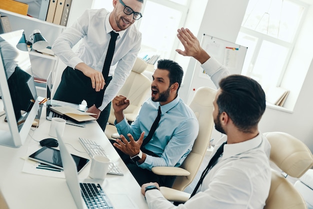 Grand succès. Groupe de jeunes hommes modernes en tenue de soirée souriant tout en travaillant au bureau