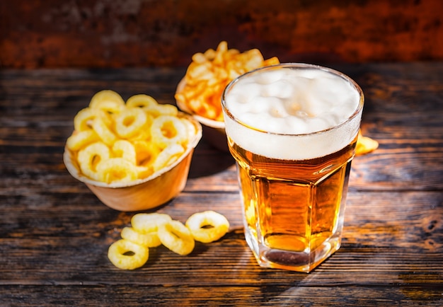 Grand verre avec de la bière légère fraîchement versée près des assiettes avec collation et frites sur un bureau en bois foncé. Concept de nourriture et de boissons