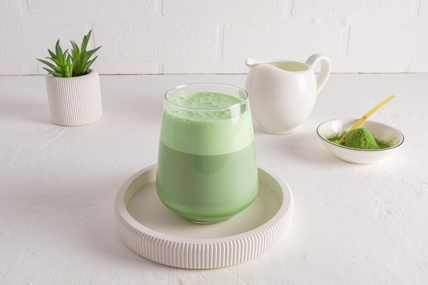 Un grand verre de thé matcha latte sain avec du lait fouetté sur un plateau rond en céramique en face d'un mur de briques blanches un matin joyeux sans café