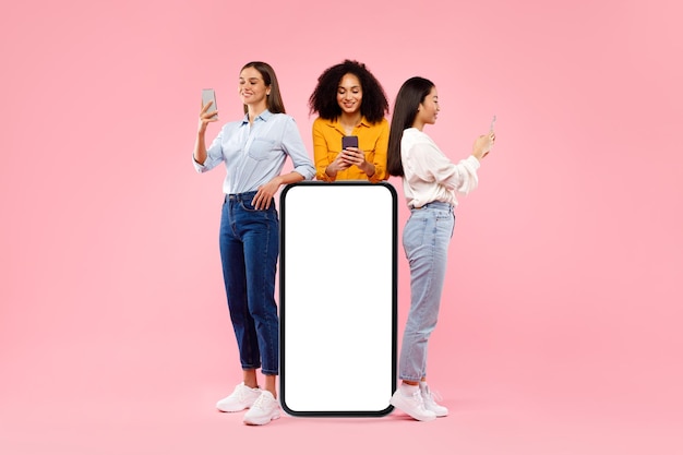 Grande application femmes multiraciales debout près d'un smartphone avec un écran blanc vierge à l'aide d'une maquette de téléphone