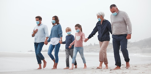 Grande famille covid et main dans la main marchant sur la plage pour un temps de liaison de qualité ensemble pendant la pandémie dans la nature Main des parents, grands-parents et enfants en liberté de voyage et promenade en famille avec des masques